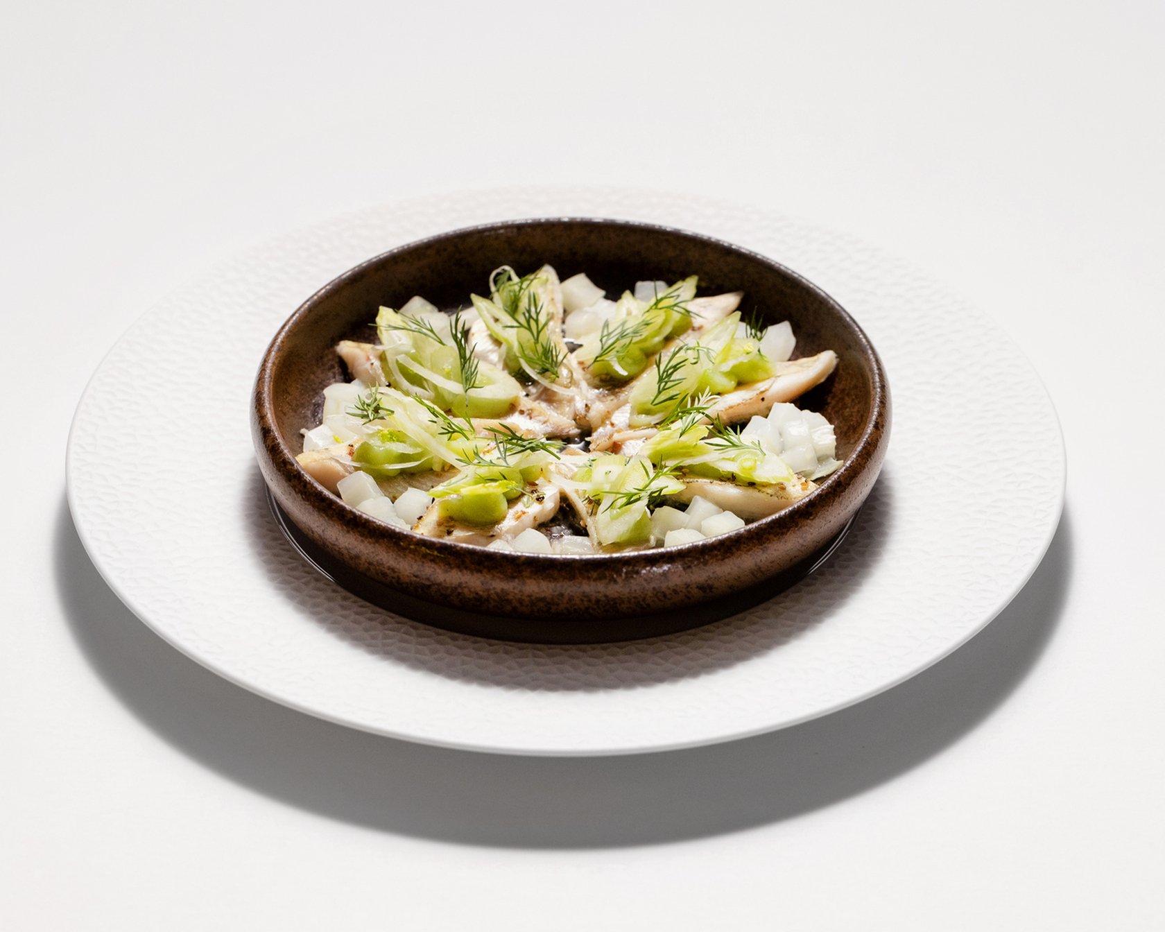 Gourmetgericht in brauner Keramikschale mit Fenchel und Kräutern auf weißem Teller